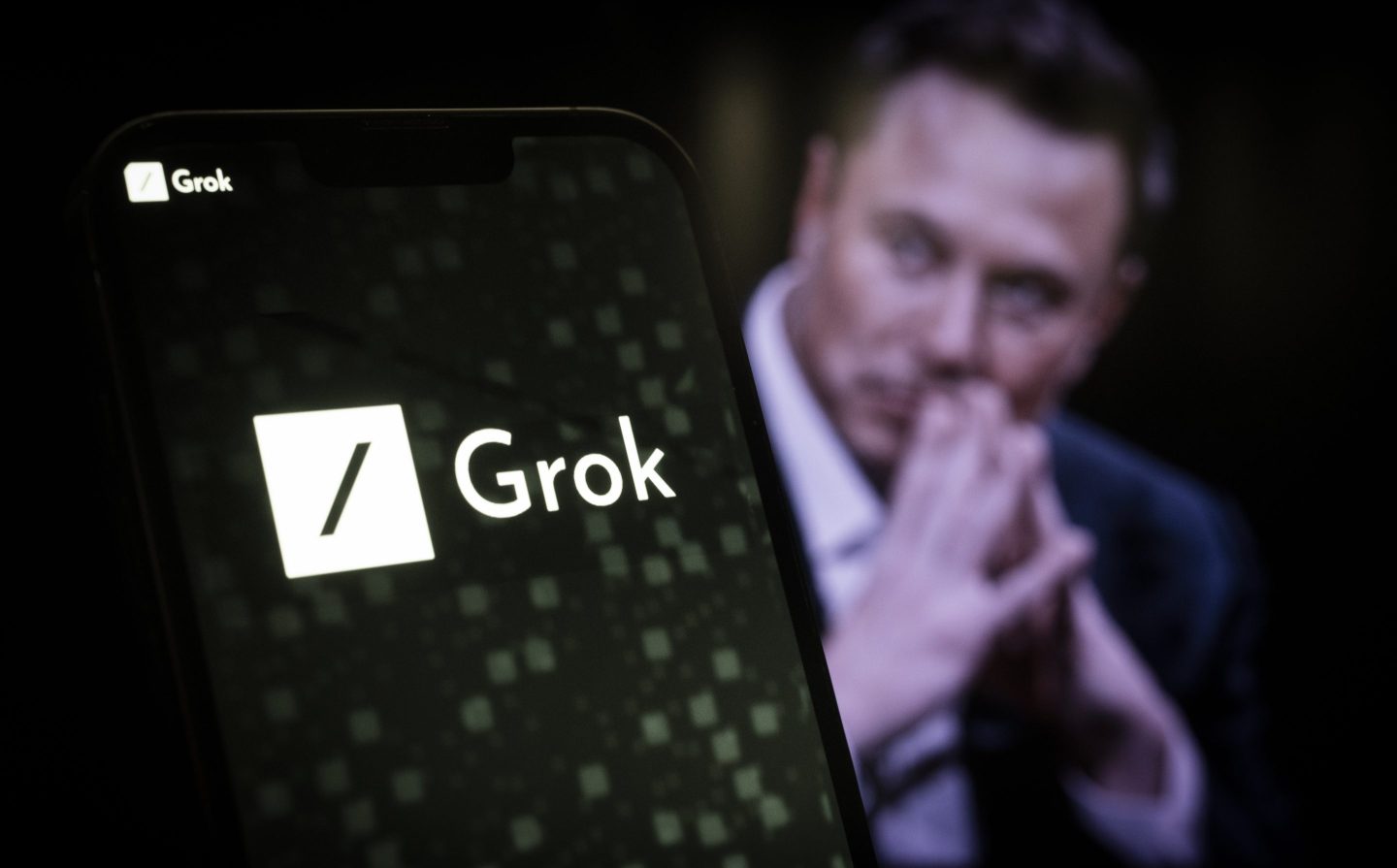 Elon Musk folds his hands behind a logo of xAI and Grok